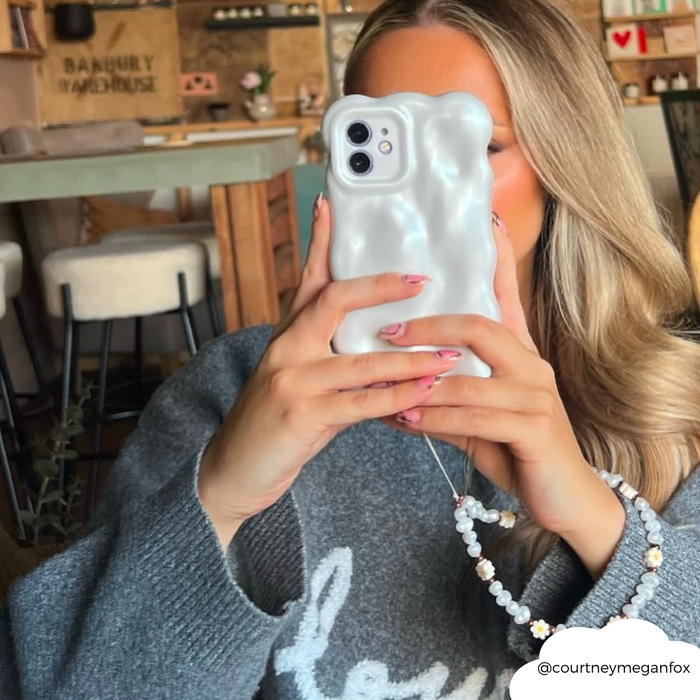 Woman holds up a white 3D pearl phone case while taking a selfie in a cozy sweater at a café.