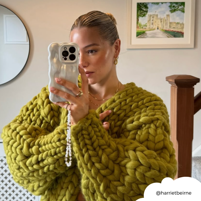Woman in green knit sweater takes a mirror selfie holding a white 3D pearl phone case; art is visible in the background.