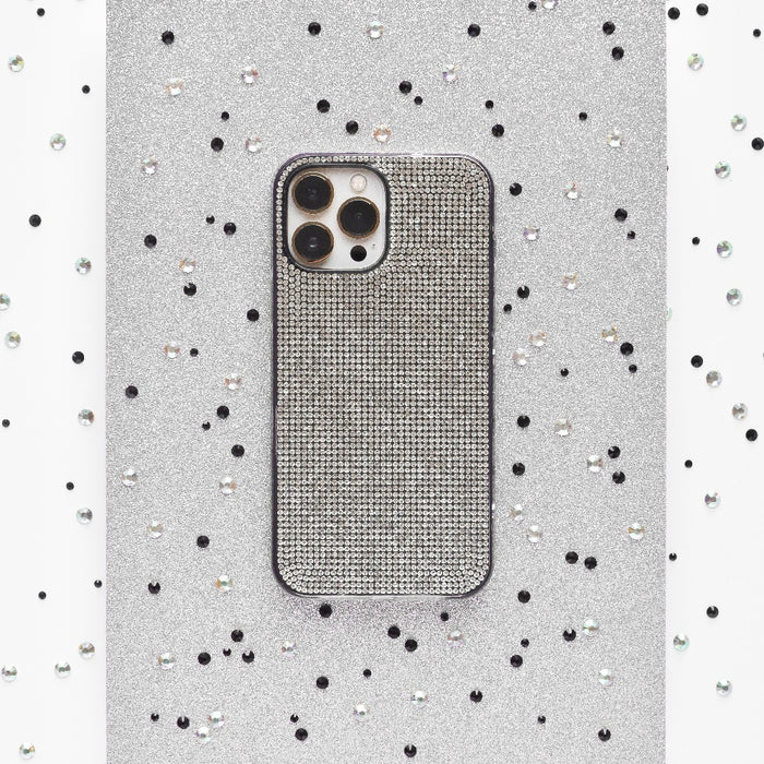 Against a sparkling silver background adorned with small black and white sequins scattered around, a smartphone is encased in the glittery Disco Sparkle Phone Case - Silver by Coconut Lane. The case features a textured, grid-like pattern and showcases three visible camera lenses on the back.
