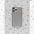 Against a sparkling silver background adorned with small black and white sequins scattered around, a smartphone is encased in the glittery Disco Sparkle Phone Case - Silver by Coconut Lane. The case features a textured, grid-like pattern and showcases three visible camera lenses on the back.