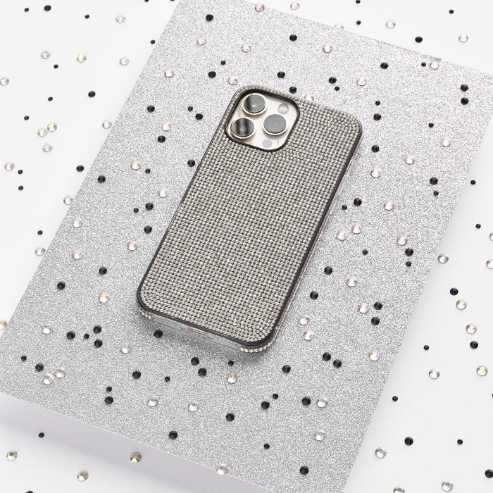 A silver iPhone adorned with the Coconut Lane Disco Sparkle Phone Case - Silver, showcasing a grid pattern, rests on a glittery silver surface. The background is sprinkled with small black and silver sequins, setting a festive and glamorous scene.