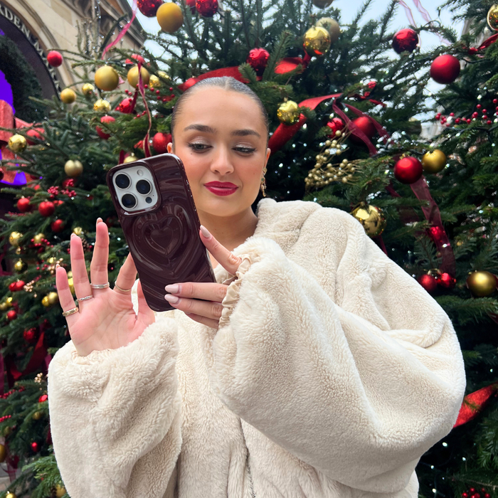 Woman in cream fur coat holds burgundy Melting Heart phone case by a decorated Christmas tree.