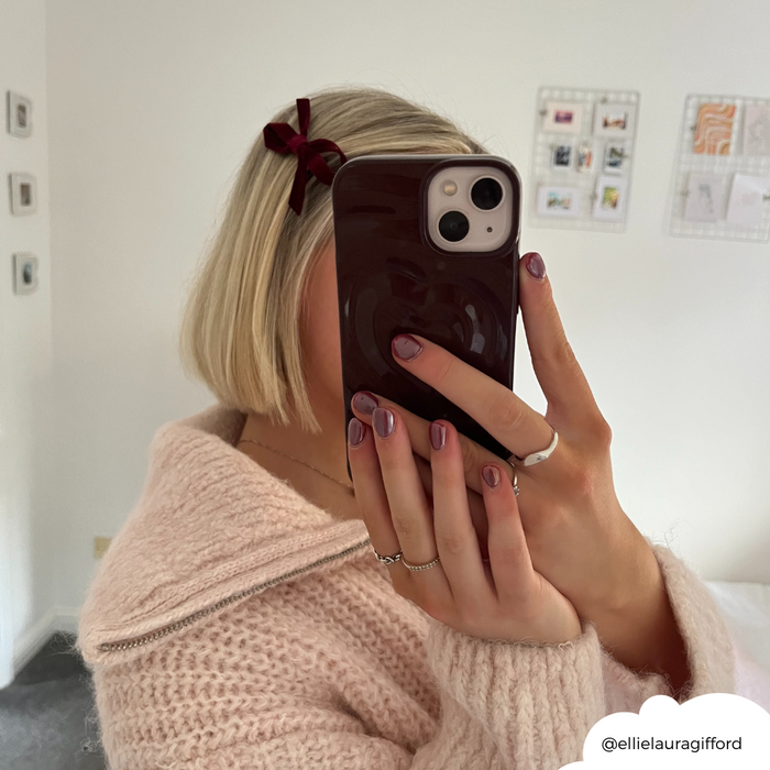 Woman with short blonde bob holds phone in burgundy Melting Heart case, showing off her purple nails in a mirror selfie.
