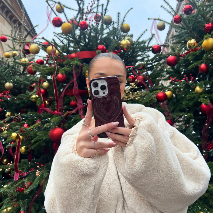 Person in beige coat takes a selfie holding a burgundy phone case with heart design, standing near Christmas trees.