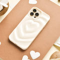 A Melting Heart Phone Case - Coconut Cream by Coconut Lane, with its smooth, pale beige case featuring a raised, 3D heart design, lies on a piece of brown paper. Surrounding the phone are small white heart-shaped cutouts and halved coconuts. The background showcases cream-colored and various brown tones.