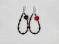 Two black beaded keychain wrist straps with heart charms — one black glossy heart, one red glossy heart — and silver swivel clips