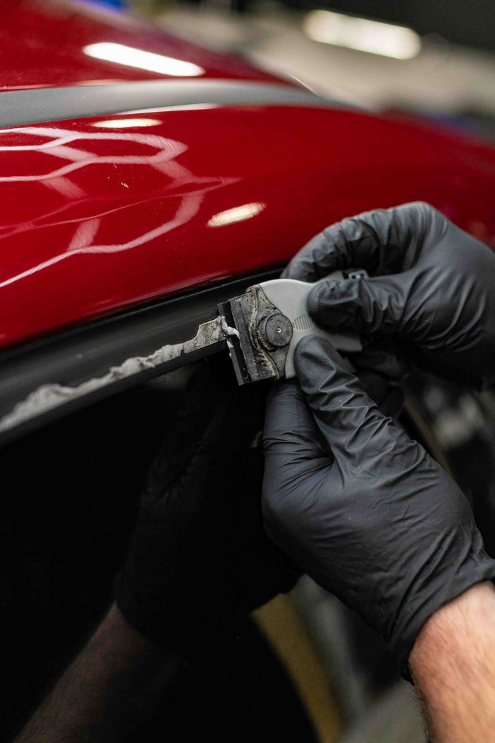 Person using a KochChemie Scraper tool on a car window of a red vehicle.