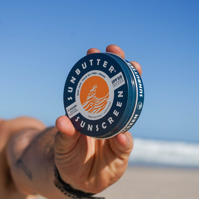 Hand holding a container of Sunbutter sunscreen with a clear blue sky background