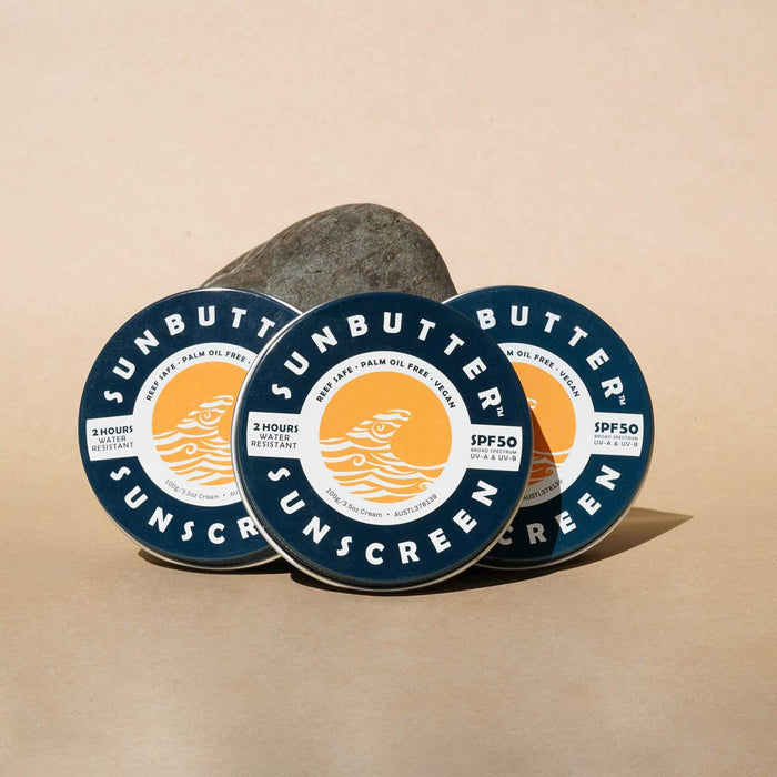 Three containers of Sunbutter sunscreen with a rock in the background on a beige surface.