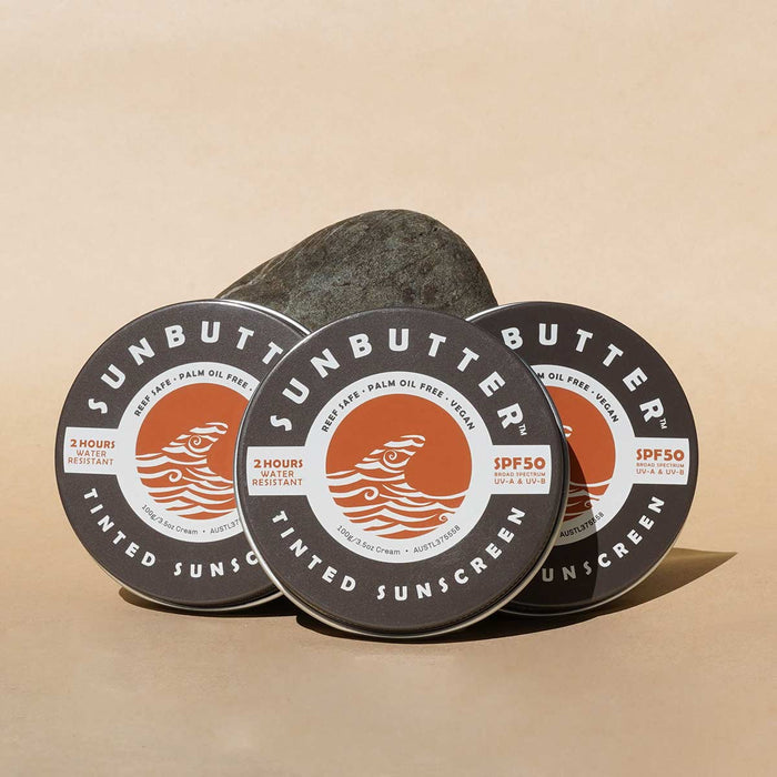 Three tins of SunButter Skincare tinted sunscreen on a beige background with a rock.
