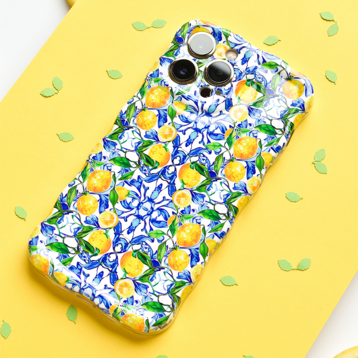 A smartphone adorned with Coconut Lane's Wavy Phone Case - Amalfi Lemon, boasting a vibrant pattern of yellow Amalfi lemons and blue flowers, is placed on a yellow surface. Green leaf-shaped confetti is scattered around the phone.