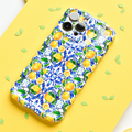 A smartphone adorned with Coconut Lane's Wavy Phone Case - Amalfi Lemon, boasting a vibrant pattern of yellow Amalfi lemons and blue flowers, is placed on a yellow surface. Green leaf-shaped confetti is scattered around the phone.